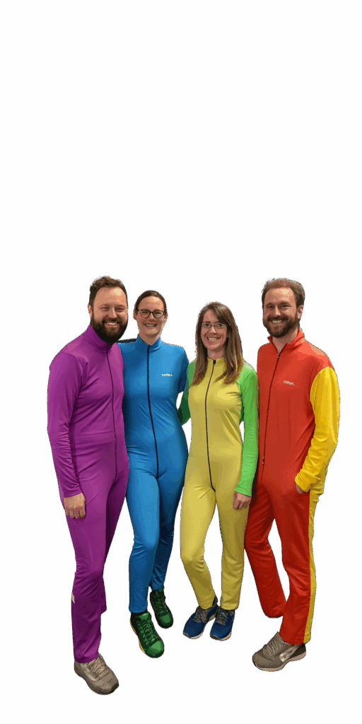 Equipo de Juegos en la Mesa posando con trajes de cuerpo completo de colores llamativos (morado, azul, amarillo y rojo), sonriendo y mostrando un estilo divertido y coordinado.