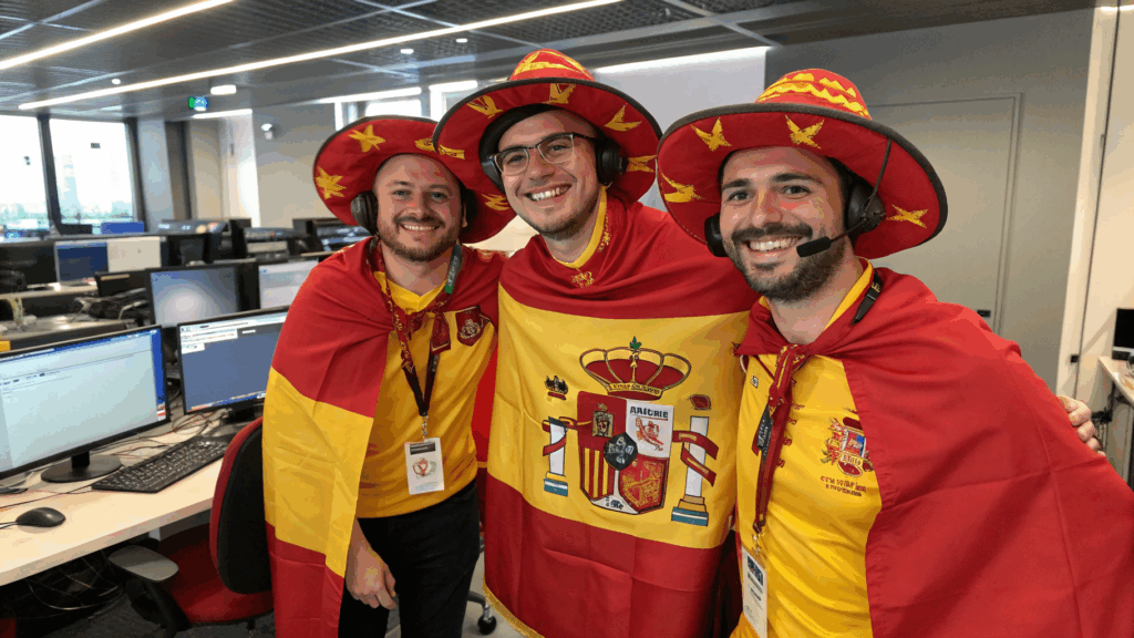 Tres hombres en una oficina vestidos con camisetas amarillas, capas con la bandera de España y sombreros rojos decorados, usando auriculares con micrófono y sonriendo, mostrando entusiasmo y espíritu de equipo.